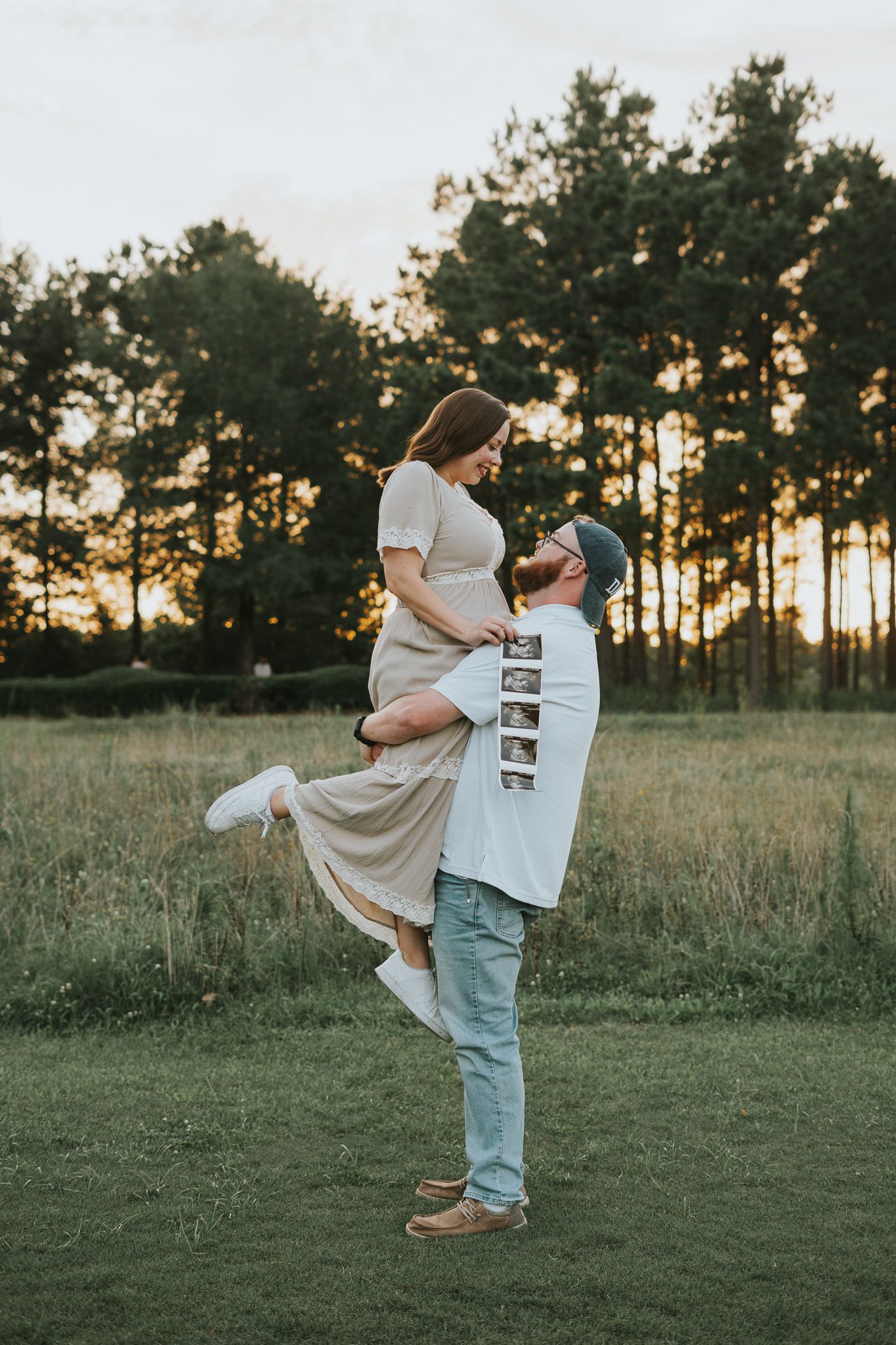 Romantic couple embracing in a field at sunset with tall pine trees silhouetted in the background.