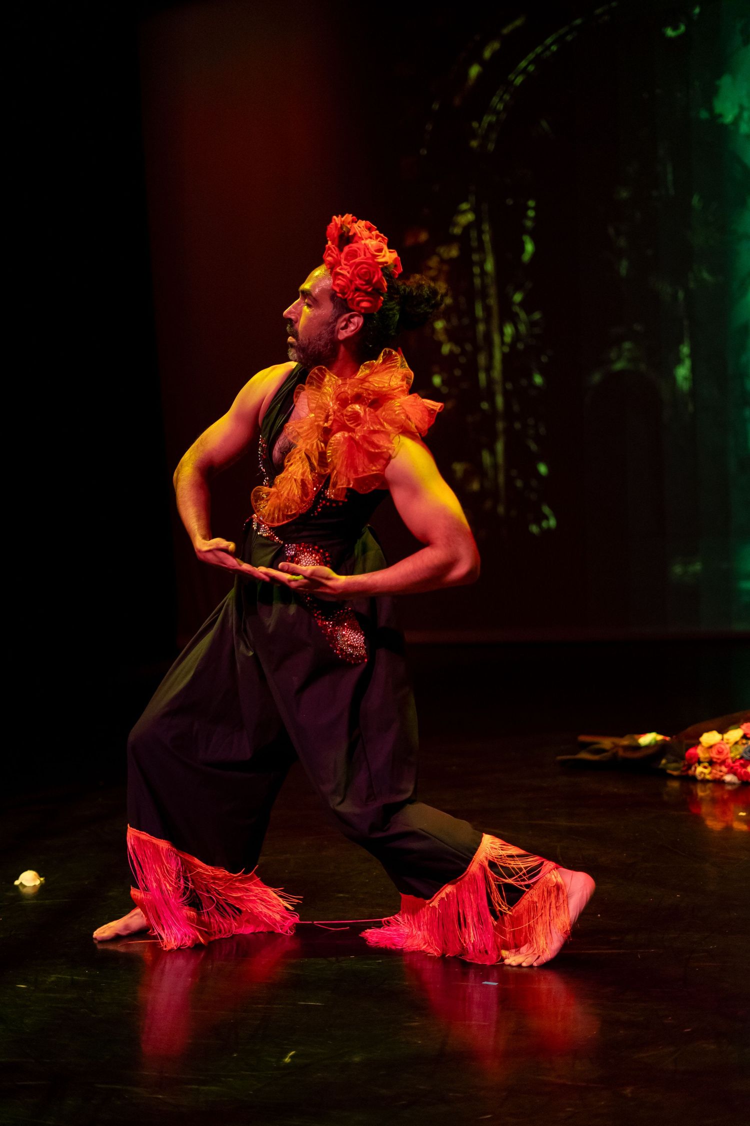 Male dancer in traditional costume with red headdress performs dramatic kneeling pose on stage under colorful lighting.