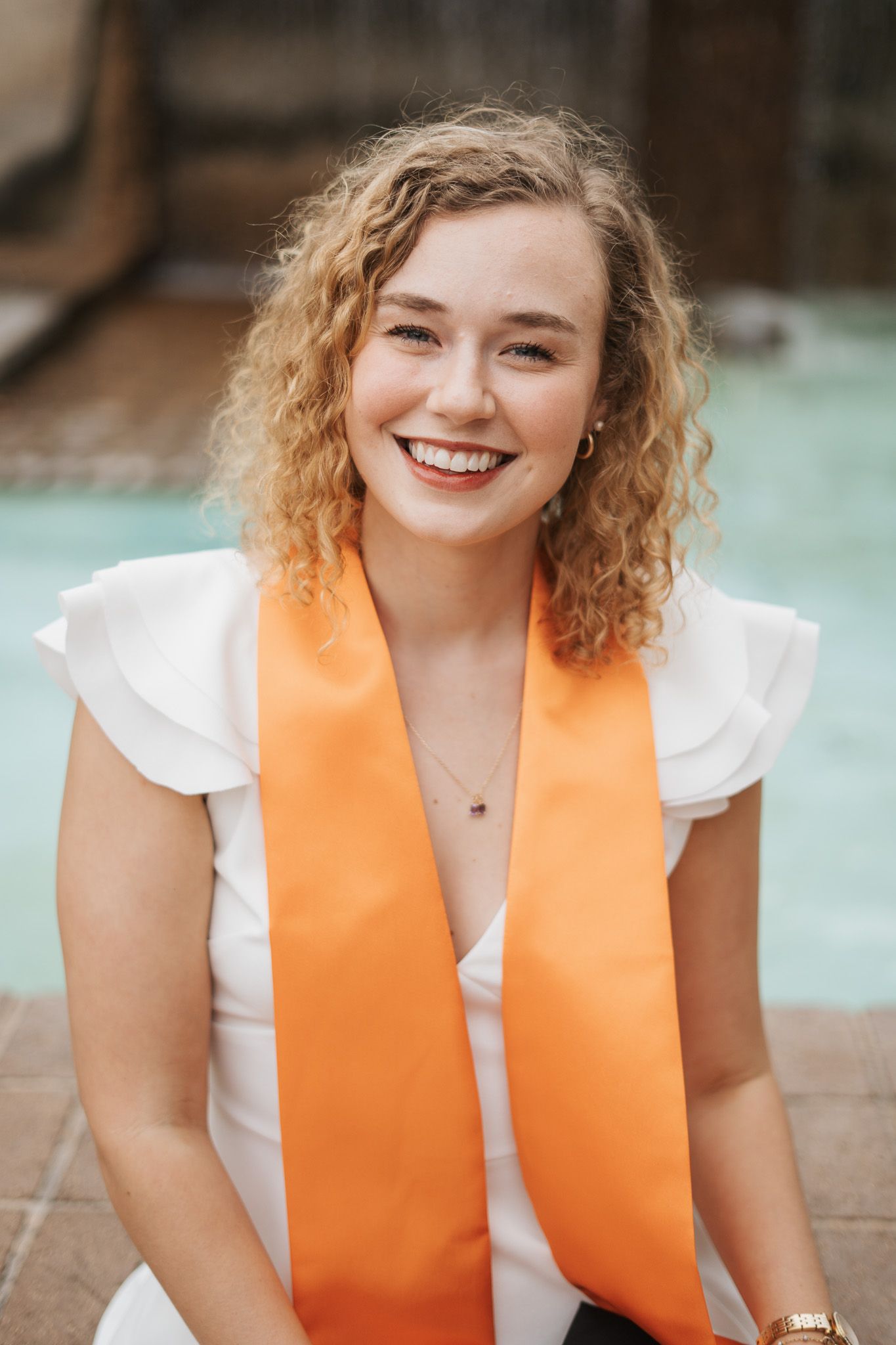 Smiling young woman wearing white top and orange graduation sash poses outdoors near a pool.