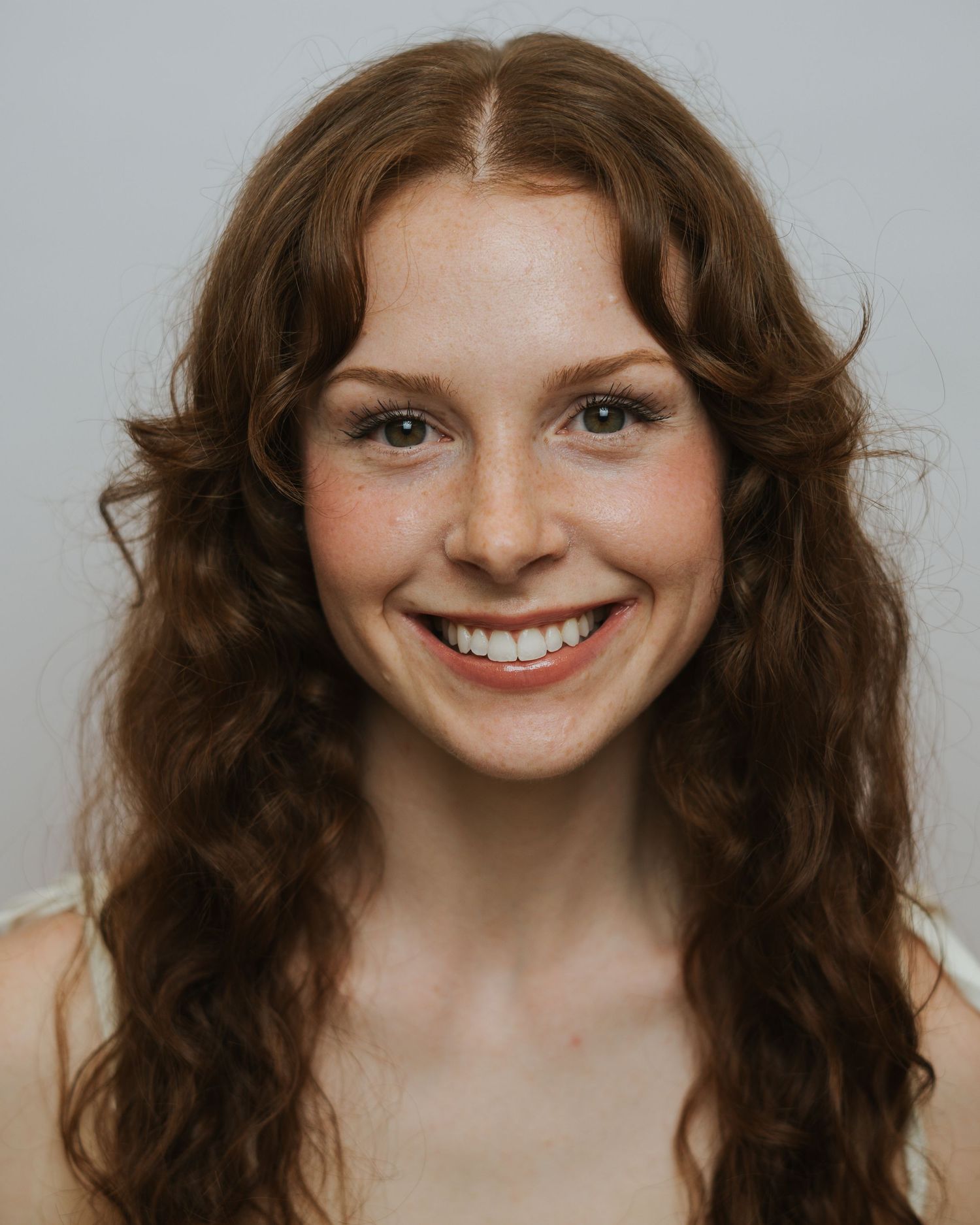 Young woman with curly auburn hair and freckles smiling warmly against a light gray background.