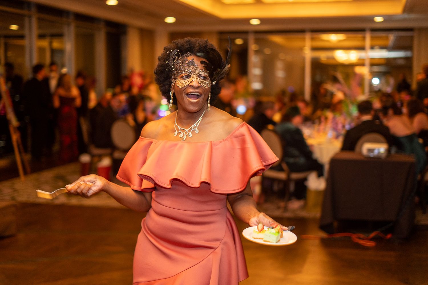 Woman in coral off-shoulder dress wearing masquerade mask dancing at elegant evening gala event.
