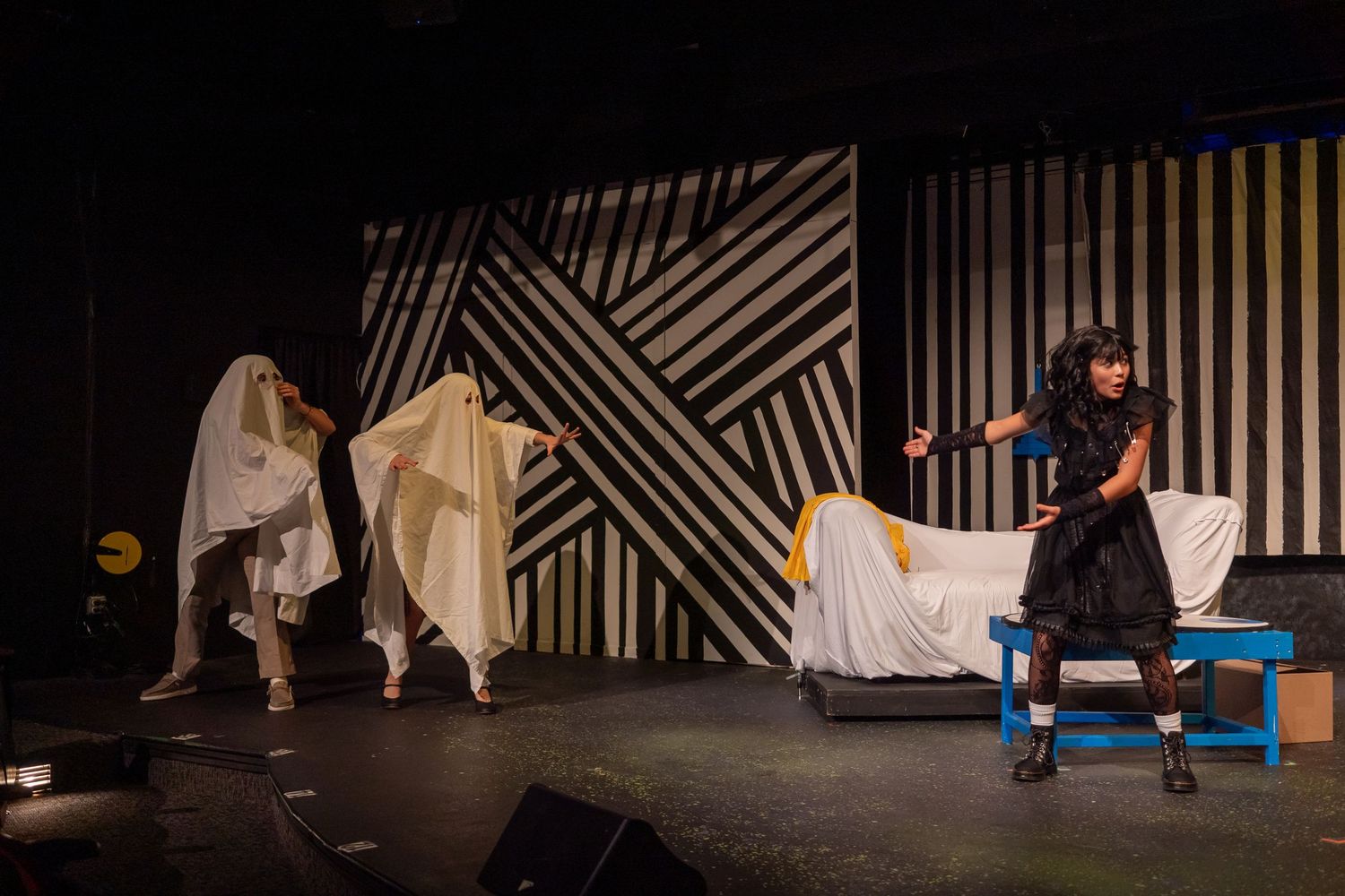 Theater performers on stage, two draped in white sheets and one in black costume, against geometric striped backdrop.