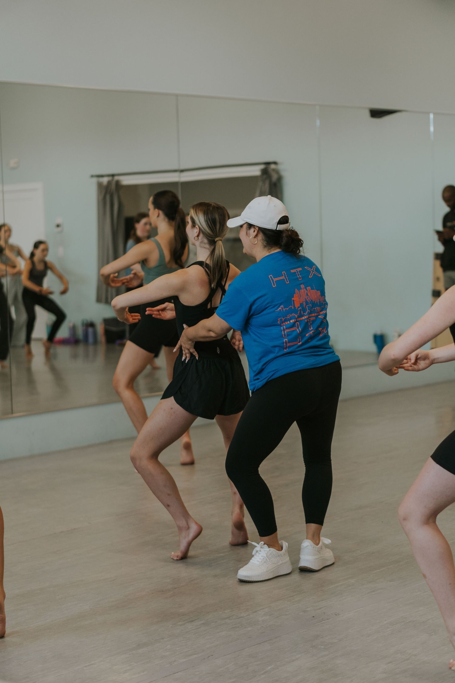 Dance class participants practicing low stance movements in a well-lit studio with mirrors.