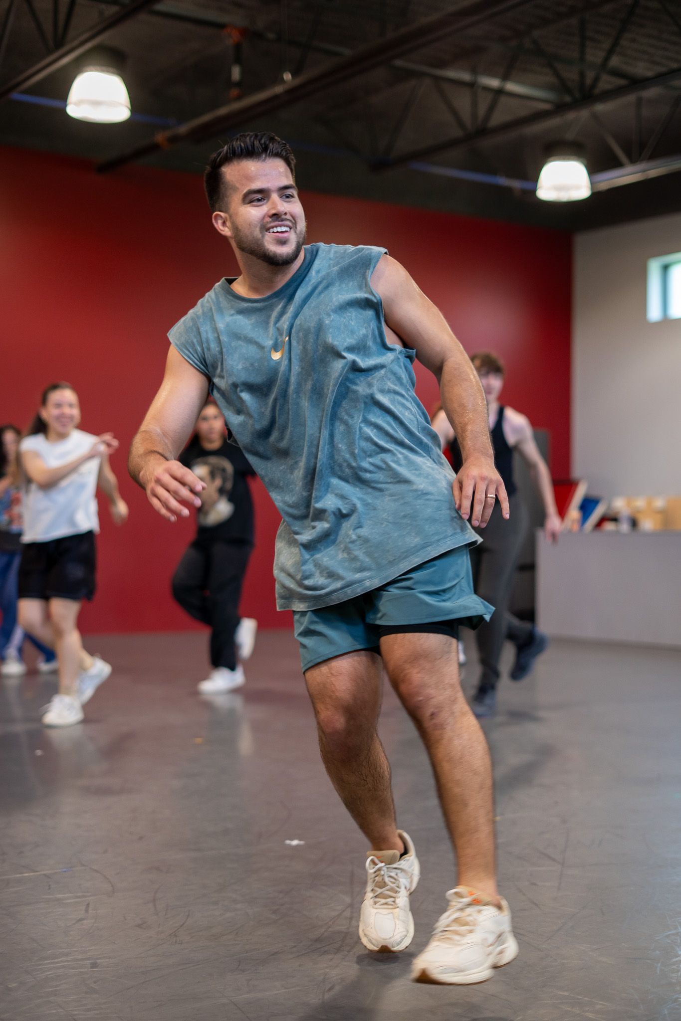 Smiling man in teal outfit dancing energetically in a dance studio with other dancers in the background.