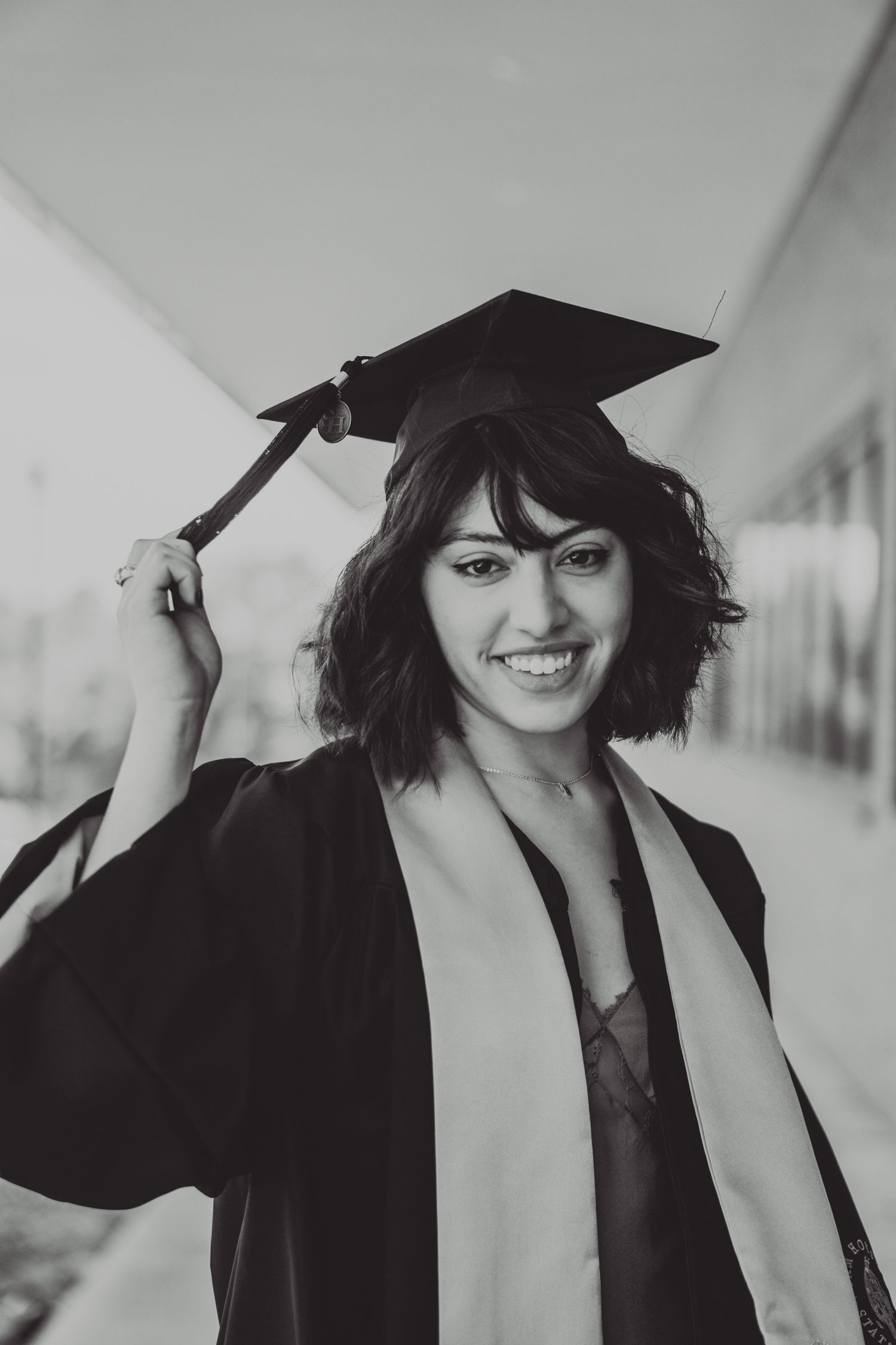 Black and white graduation portrait of graduate adjusting mortarboard cap with bright smile.