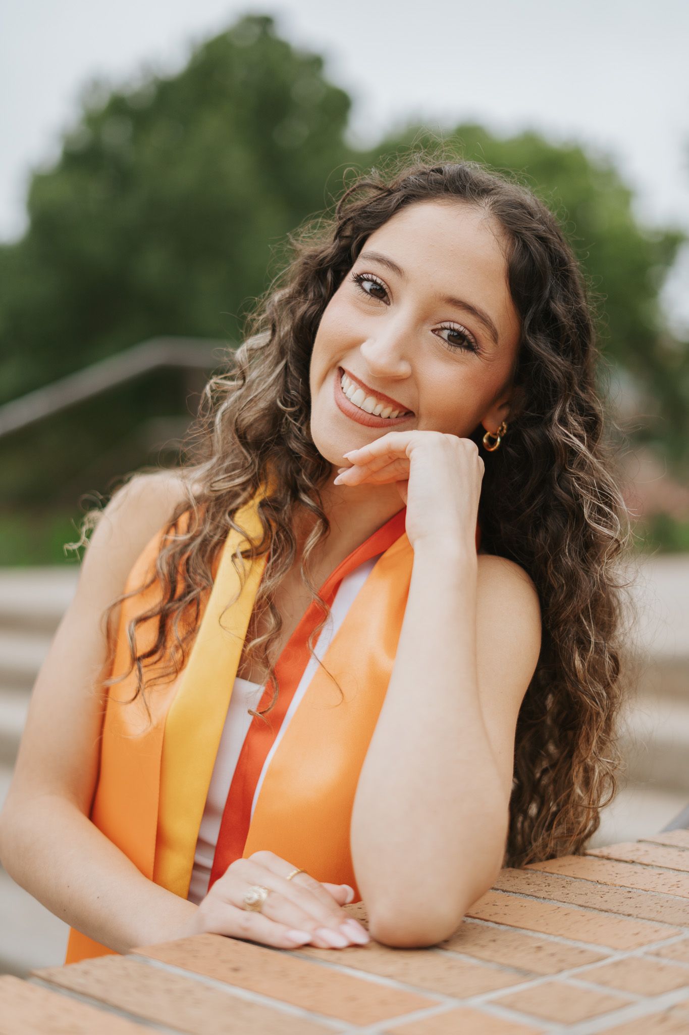 Smiling young woman in orange graduation regalia with curly hair poses outdoors on steps.