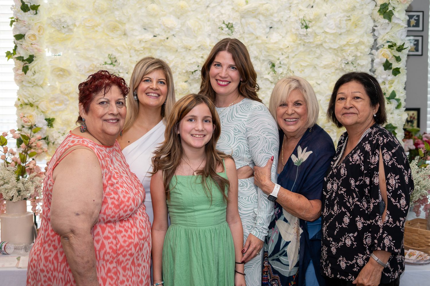Six women and a young girl pose together smiling in front of a white floral wall backdrop at a celebratory event.
