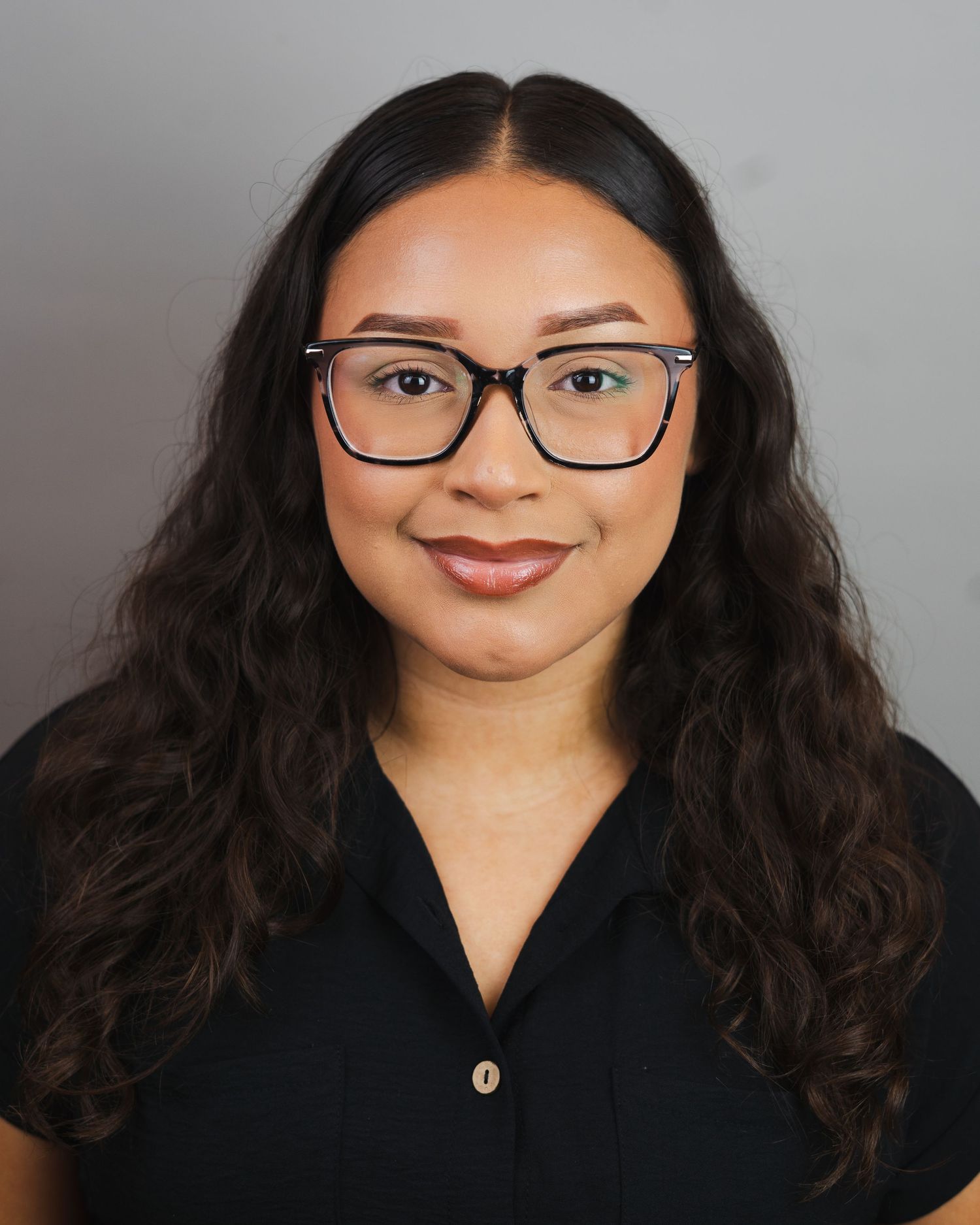 Professional headshot of a young woman with long dark curly hair, glasses, and a black top against a gray background.