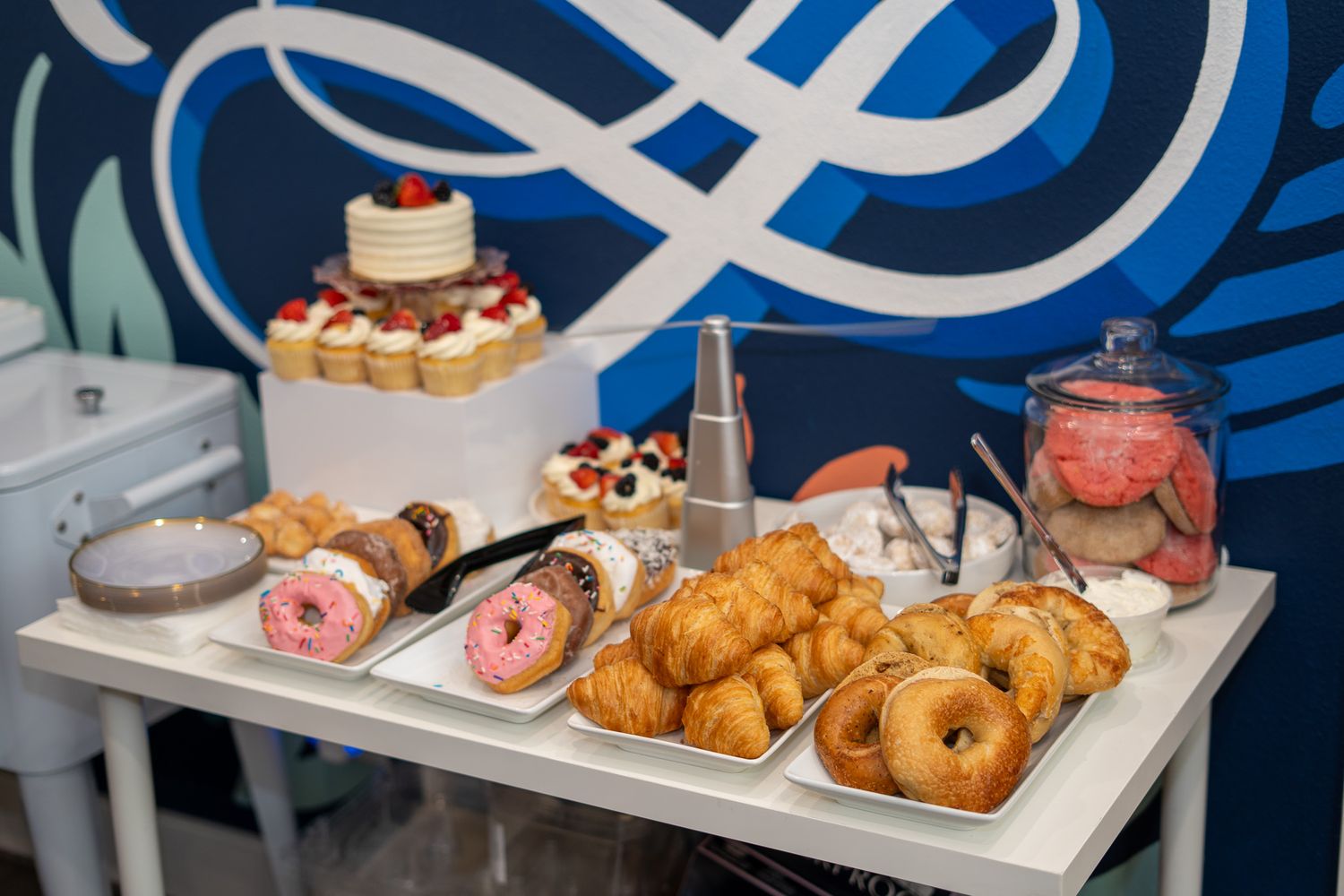 A dessert table with donuts, croissants, and a layered cake displayed on a white cart against a blue wall.