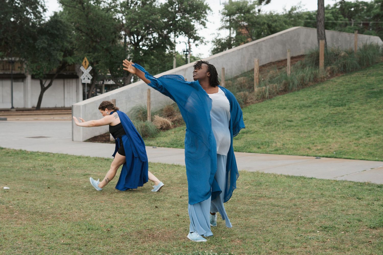 Two dancers perform outdoors in flowing blue garments on a grassy area near a street.