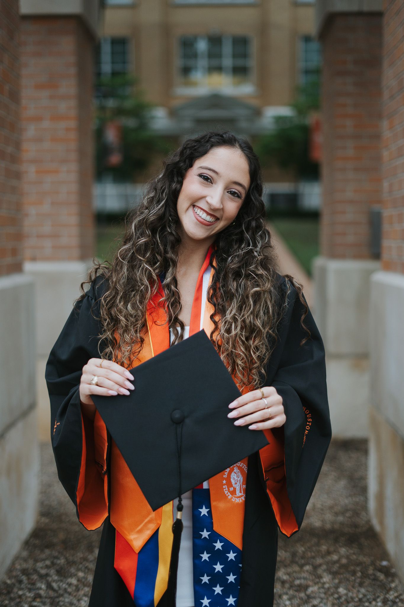 Smiling graduate in black gown holding cap, wearing orange stole and American flag sash outside brick building.