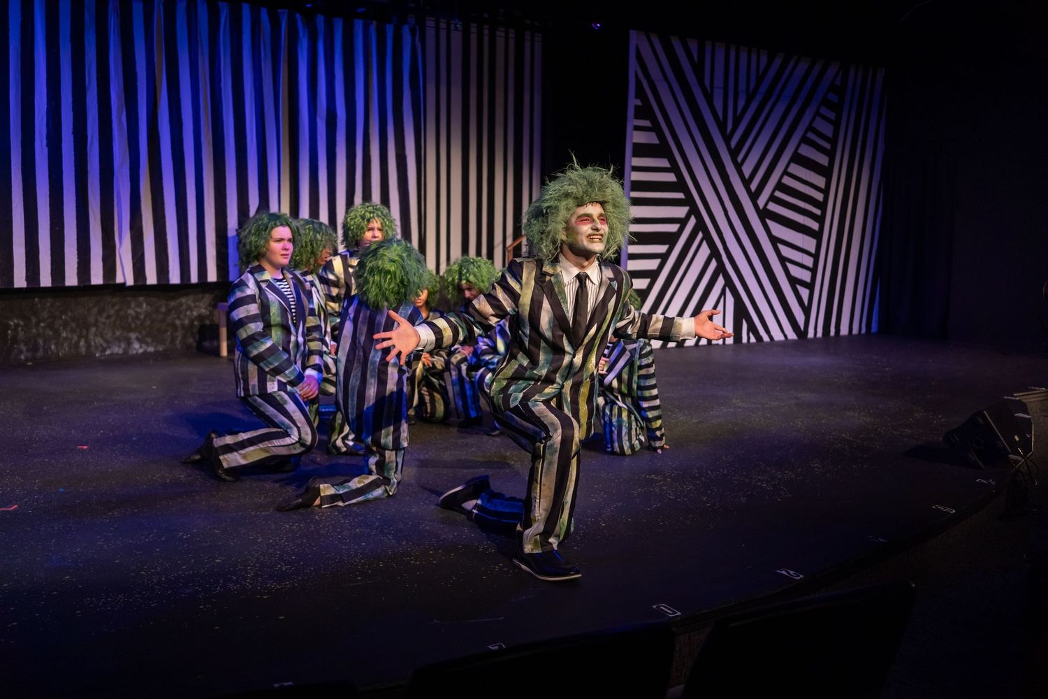 Beetlejuice stage musical performers in striped costumes with green hair on a theatrical set.