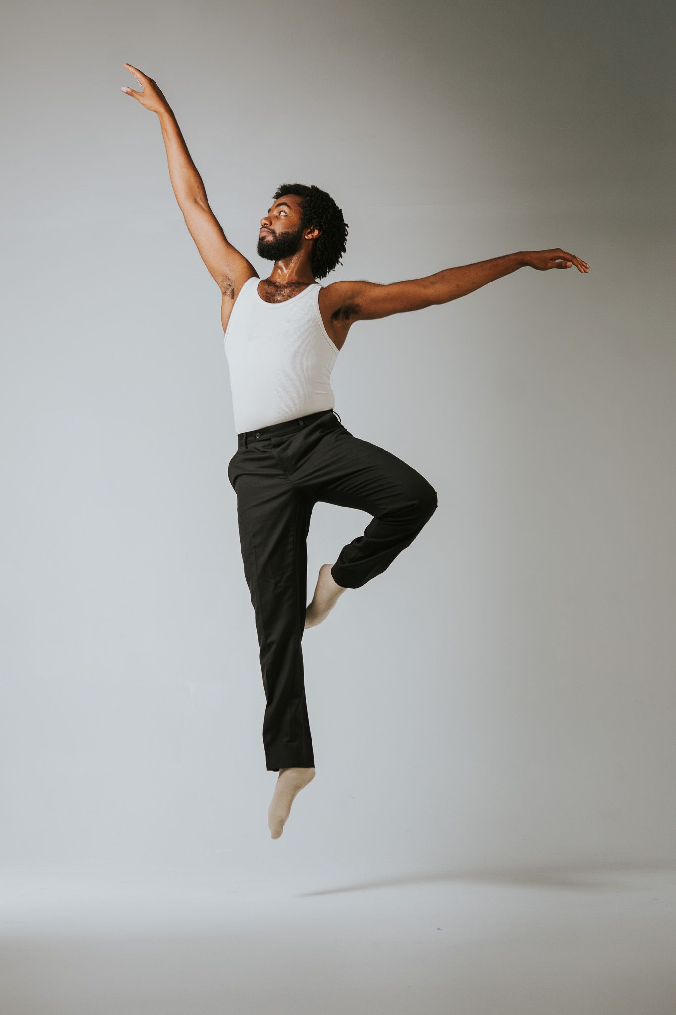 Modern dancer performs expressive leaps in white tank top and dark pants against light background.