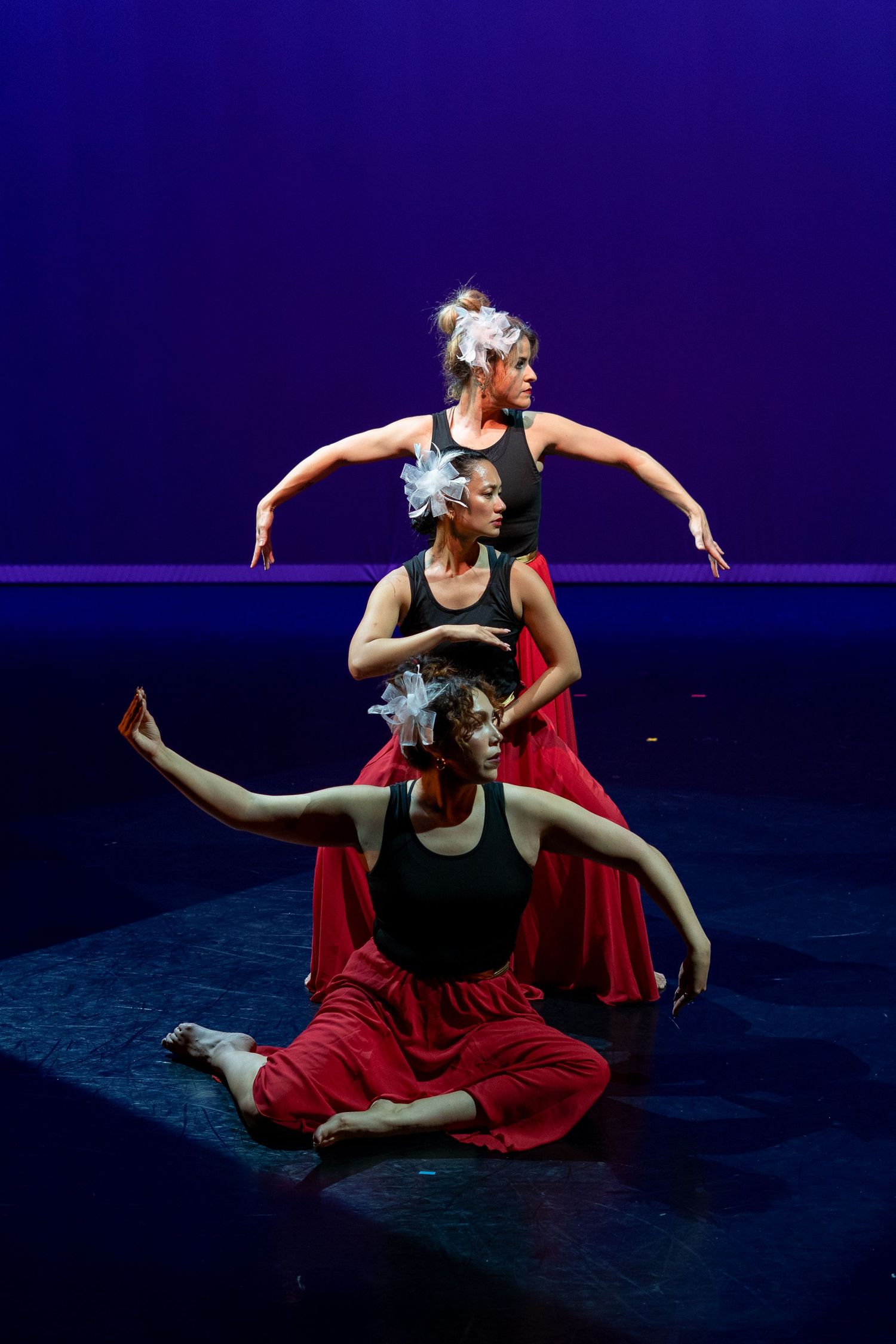 Three female dancers perform on stage in black tops and red skirts under blue and purple lighting.