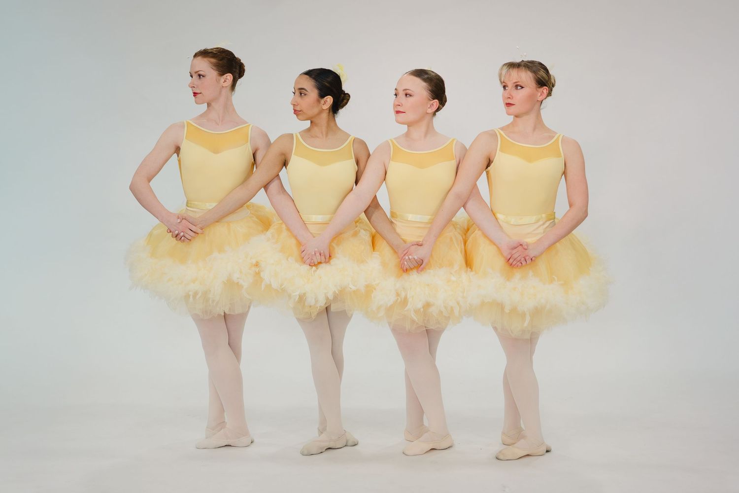 Four young ballerinas in matching yellow tutus and ballet shoes pose gracefully in a studio setting.
