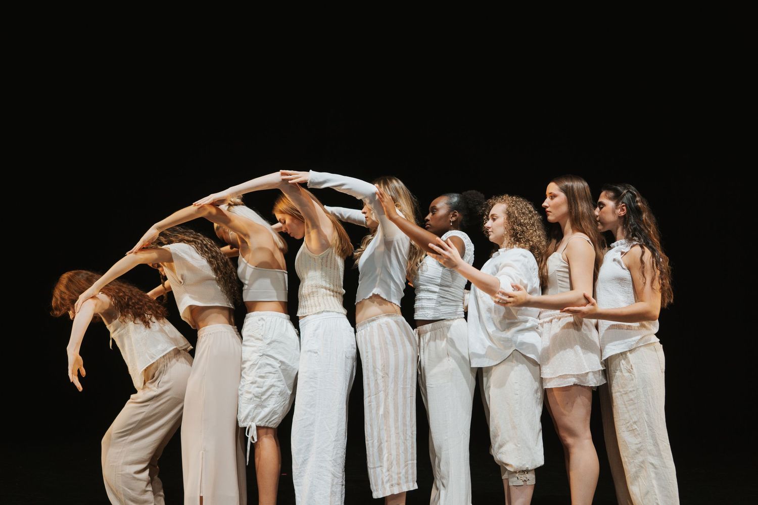 Group of female dancers in white outfits performing a contemporary dance routine against a black background.