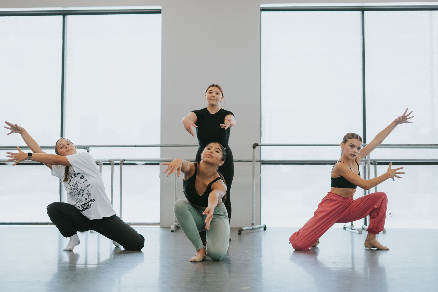 Dancers strike dynamic poses in a bright dance studio with floor-to-ceiling windows.