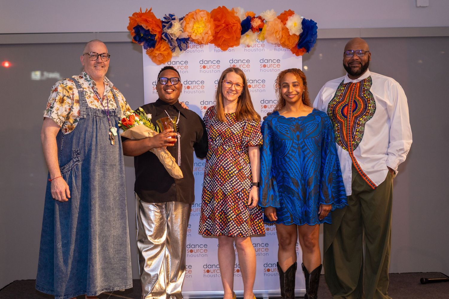 Five people pose together at a colorful event with orange and blue floral decorations, wearing vibrant cultural outfits.