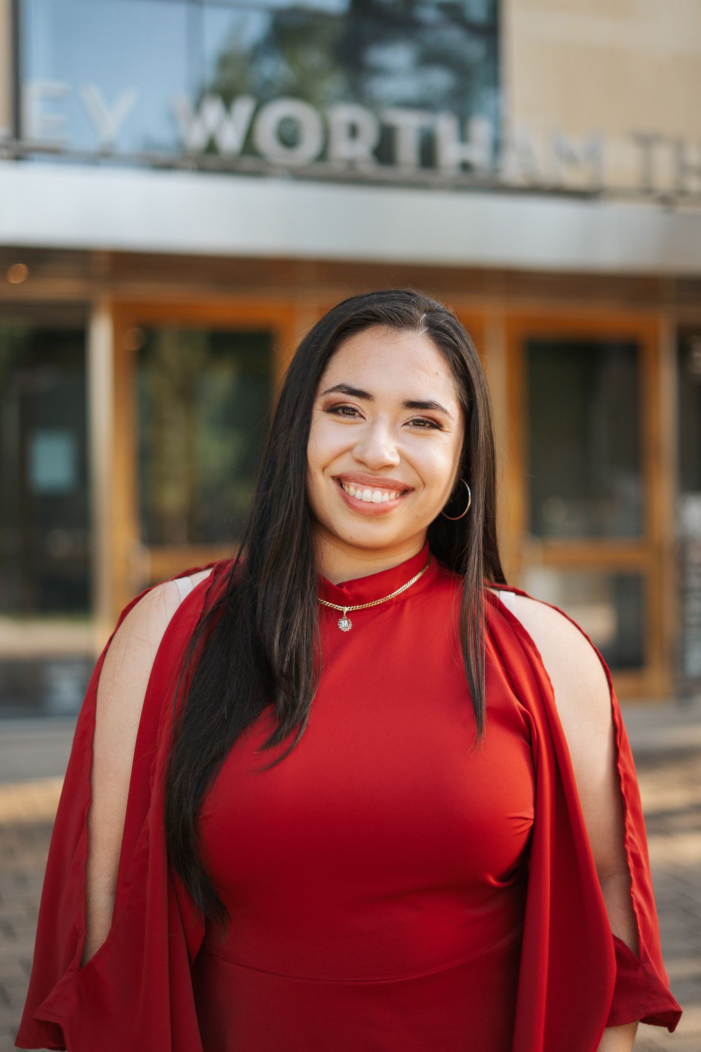 Smiling young woman in red dress and cape standing outdoors near a building entrance.