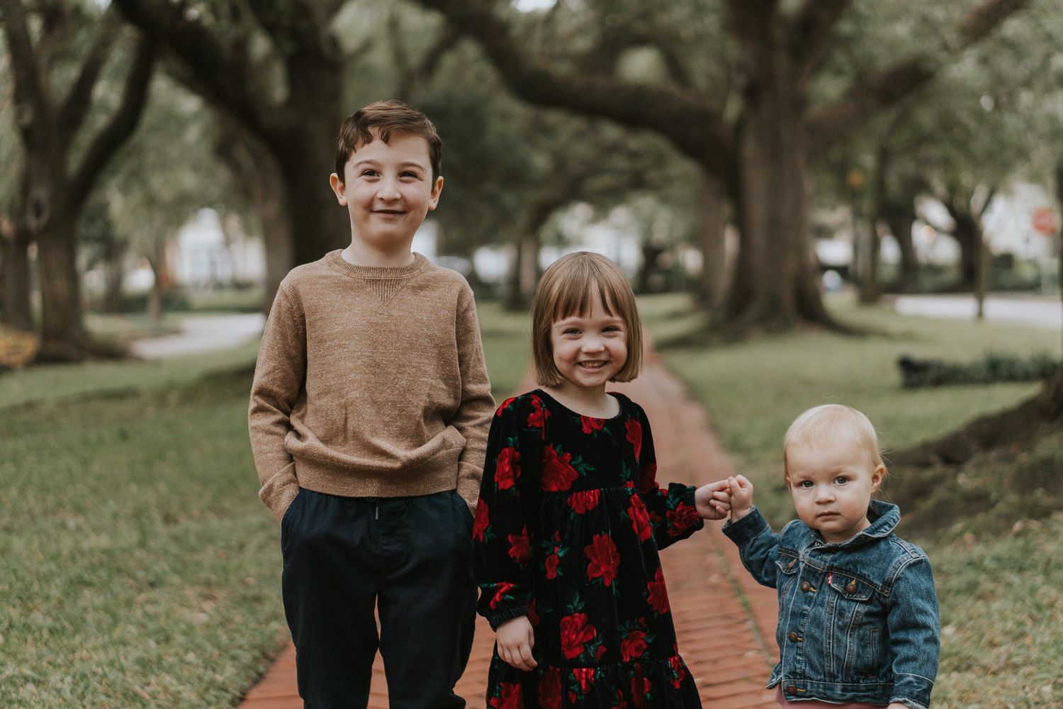 Three young children standing together on a brick pathway under oak trees.