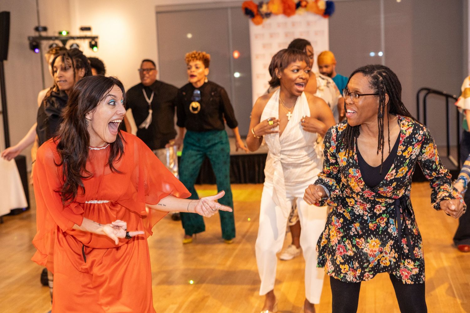 Women dancing joyfully at a festive indoor event on a wooden dance floor.