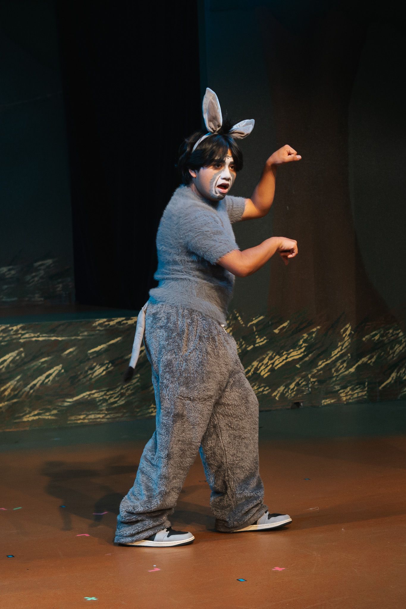 A performer in a gray donkey costume with ears and face paint dances on a theatrical stage.