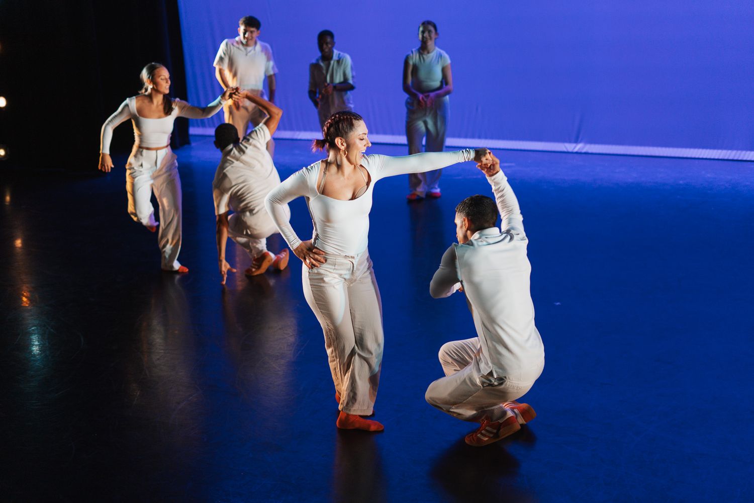 Ballet dancers perform synchronized choreography on a stage illuminated by dramatic blue lighting while wearing white costumes.