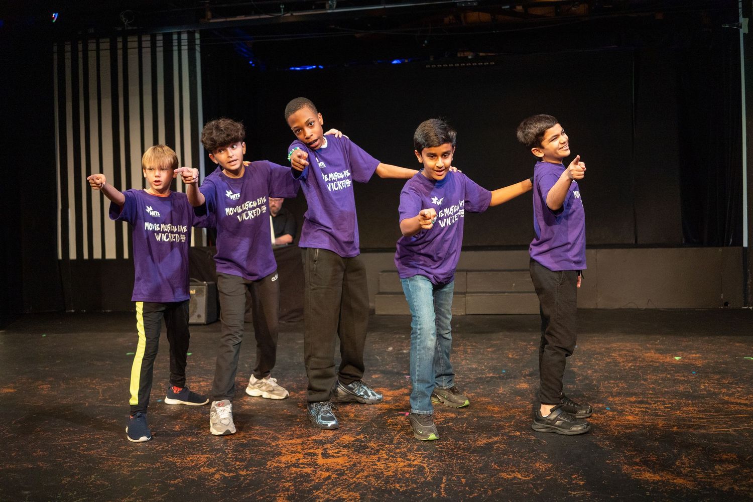 Five boys in purple matching t-shirts perform a dance routine on stage under bright lights.