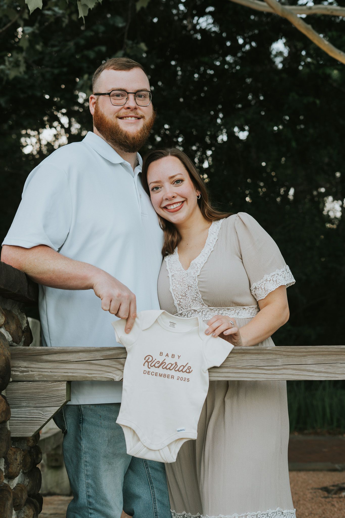 Expectant couple holding a baby onesie outdoors during a pregnancy photoshoot.
