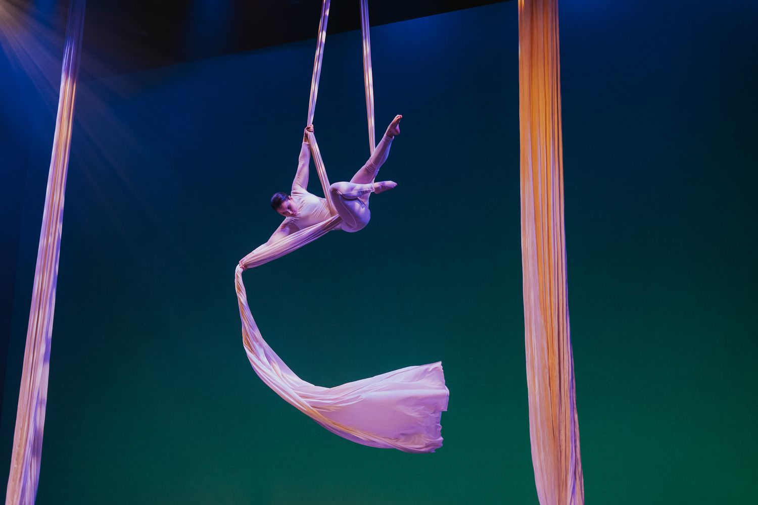 An aerial silk performer creates graceful shapes with purple fabric against a blue-green backdrop.