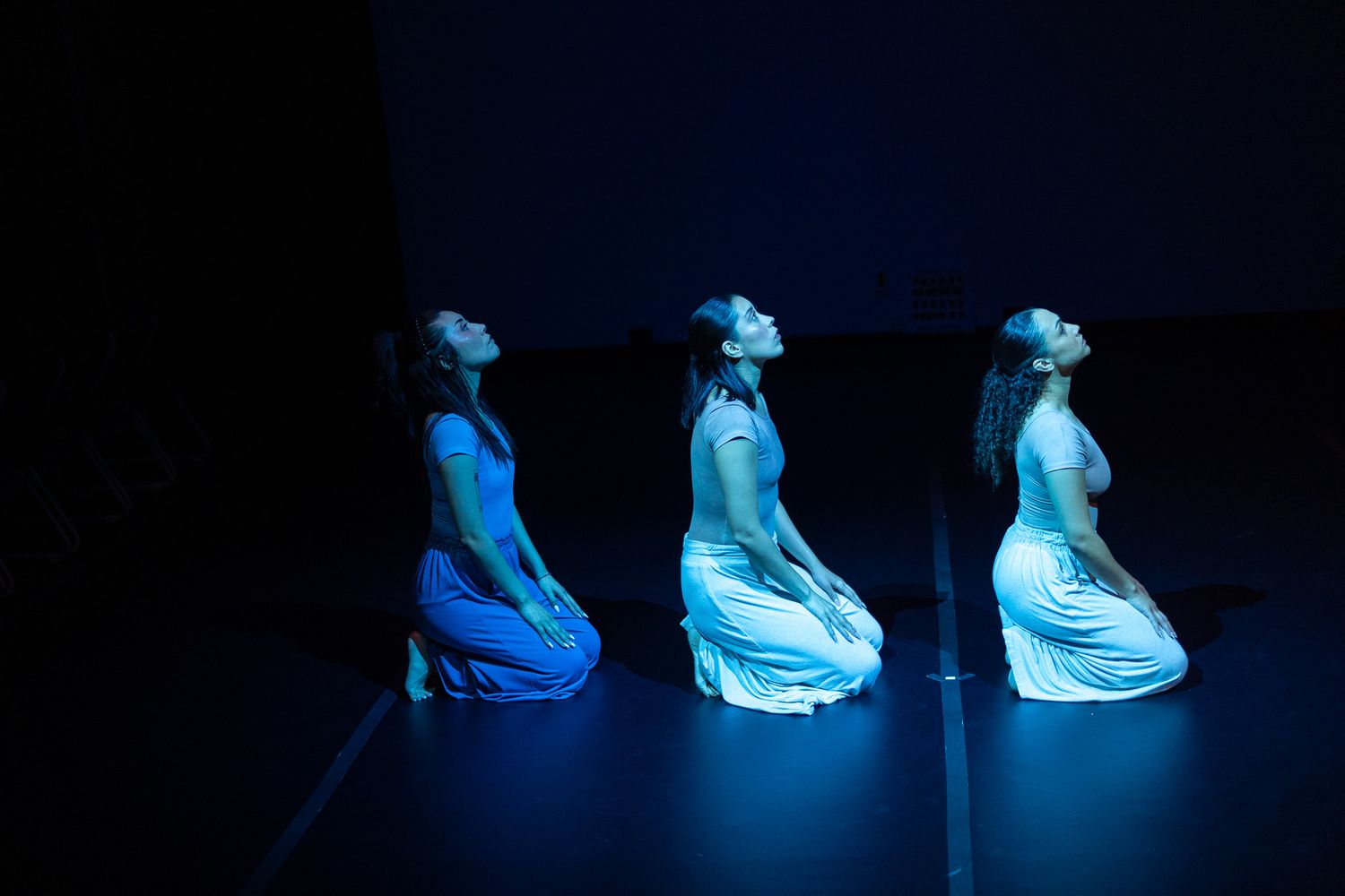 Three dancers in white dresses kneel on a dark stage, illuminated by blue light, heads tilted back gracefully.