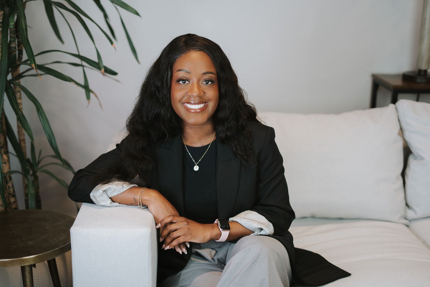 Smiling young Black woman in black blazer and gray pants sitting on white couch beside indoor plant.