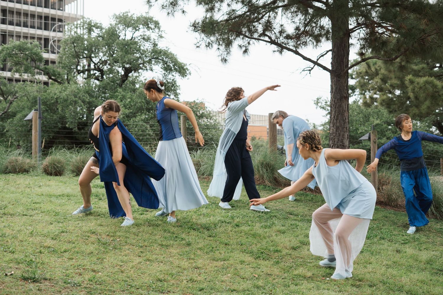 Dancers in blue and white costumes perform an outdoor contemporary dance routine on a green lawn with trees and a building.