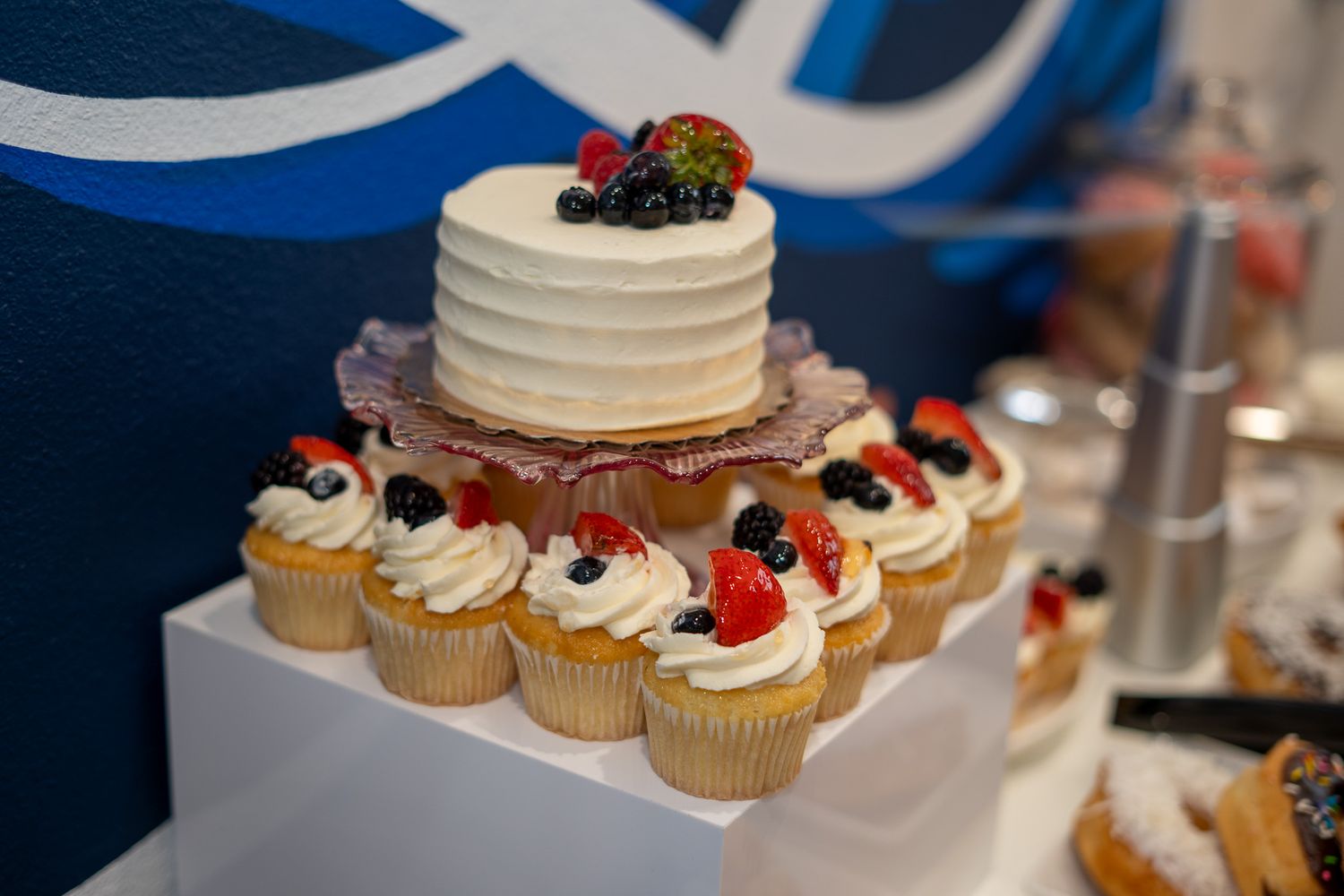 White frosted cake topped with fresh berries on a tiered stand surrounded by matching berry-topped cupcakes.
