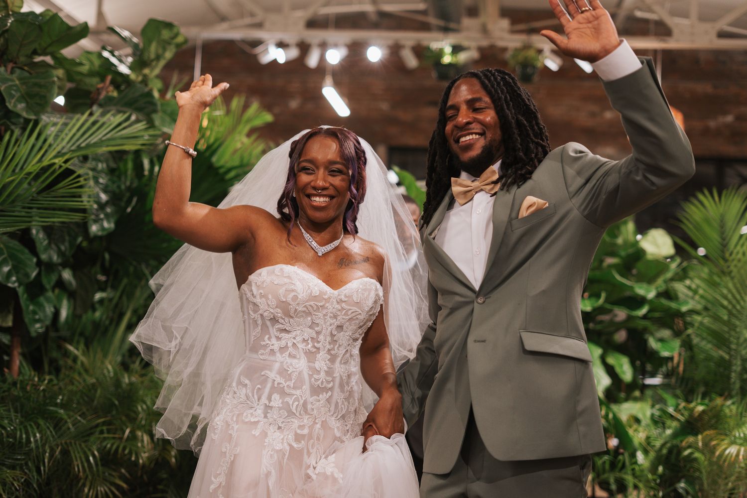 Joyful newlywed couple celebrating in lush botanical venue, bride in lace gown and groom in gray suit.
