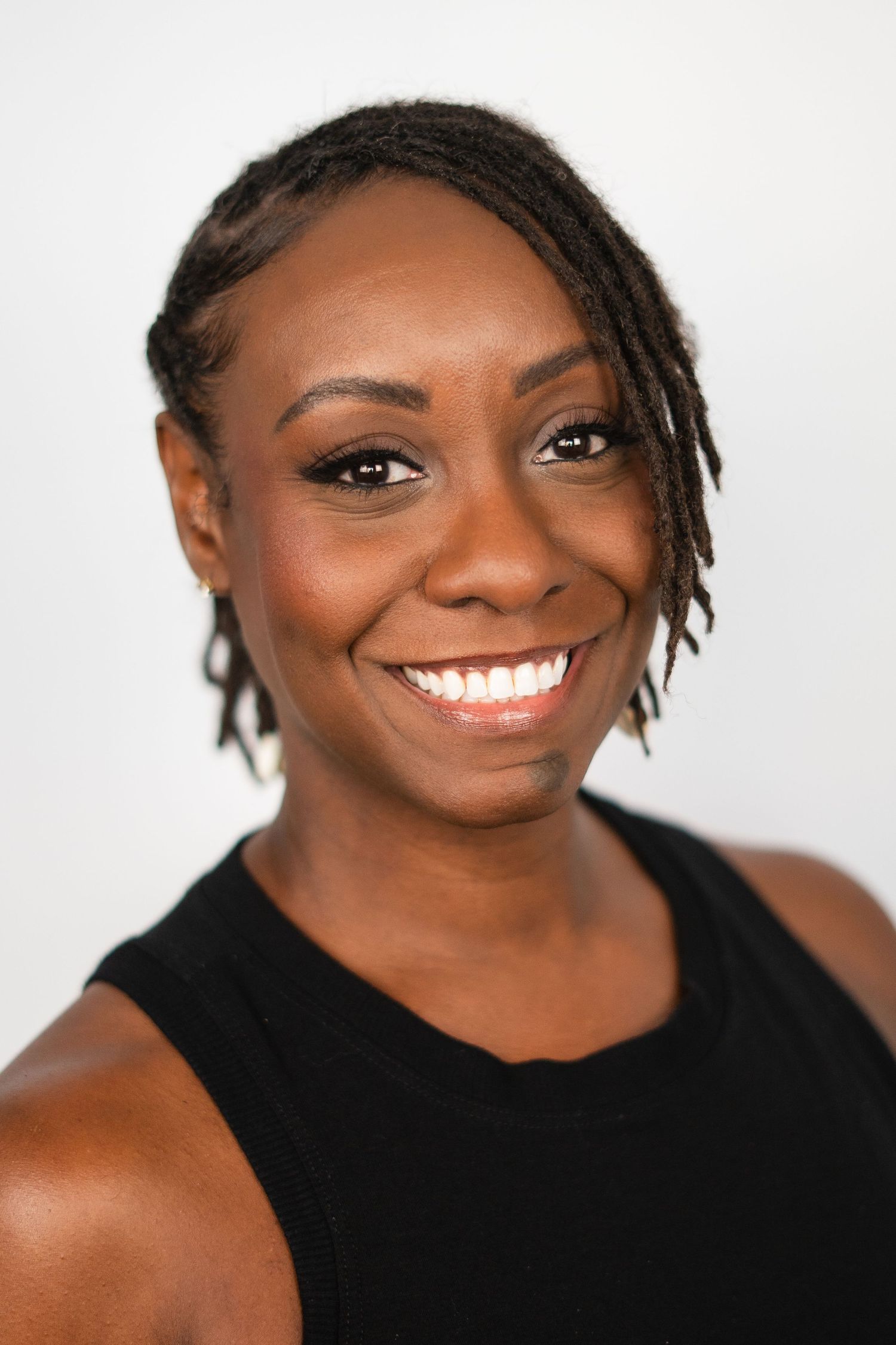 Smiling Black woman with short dreadlocks wearing a black sleeveless top against a white background.