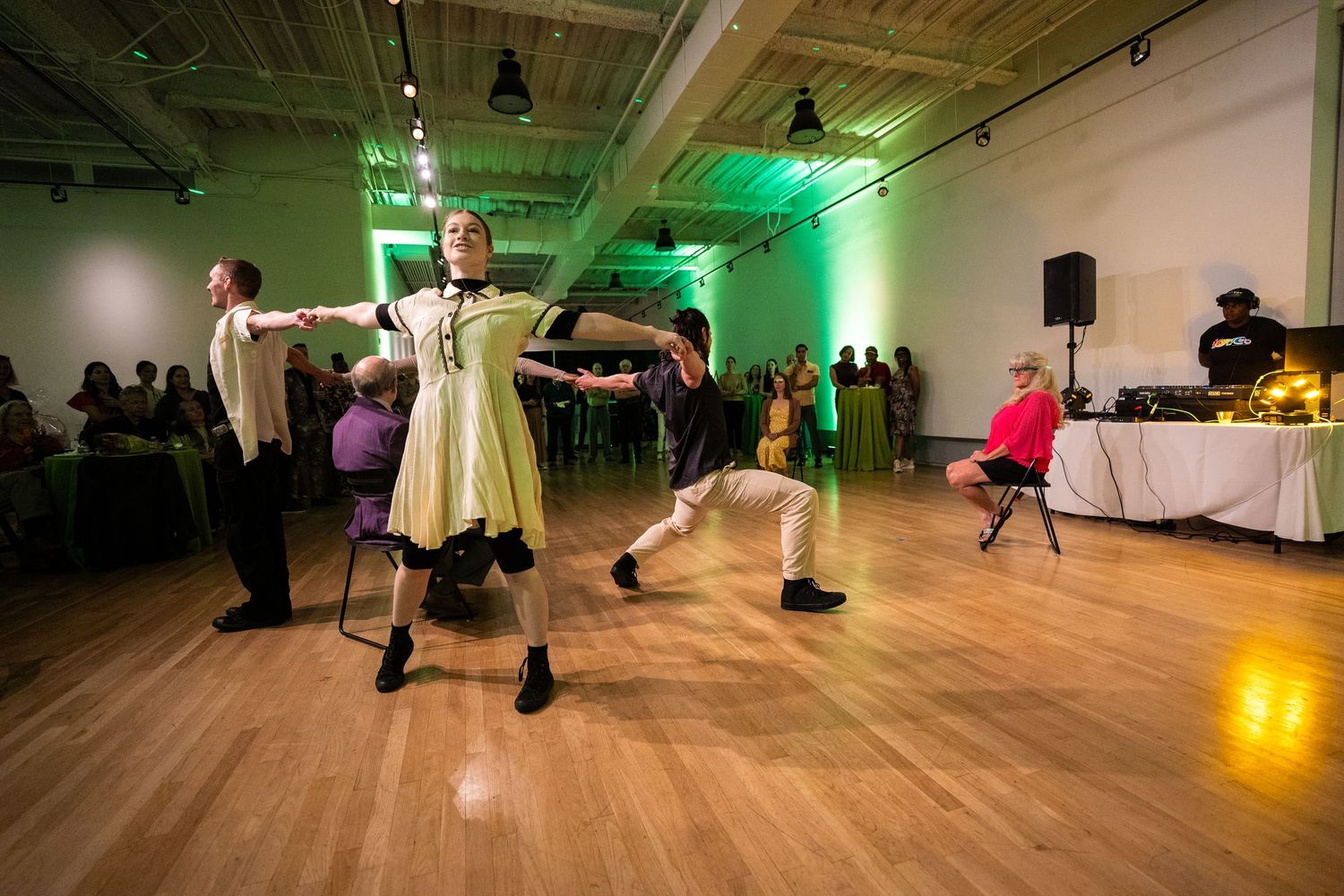 Dancers perform energetic moves on a wooden dance floor with green ambient lighting at a social gathering.