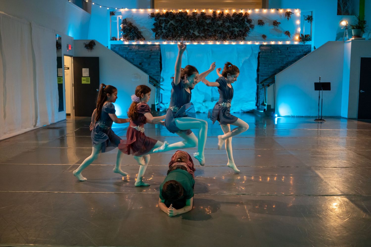 Young girls perform a dynamic dance routine on a stage with blue lighting and marquee lights in the background.