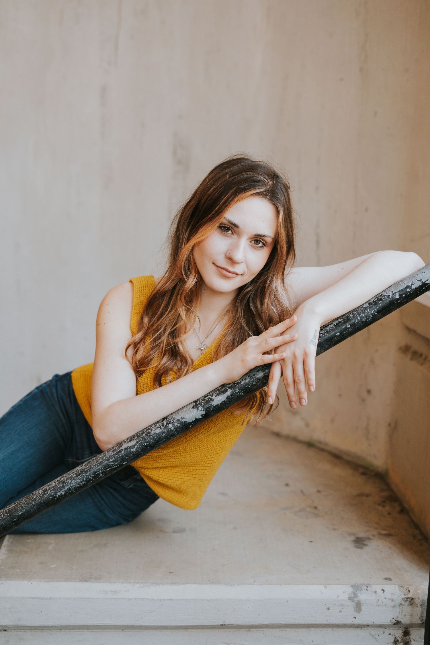 Portrait of woman in yellow tank top and jeans leaning on railing against light wall.