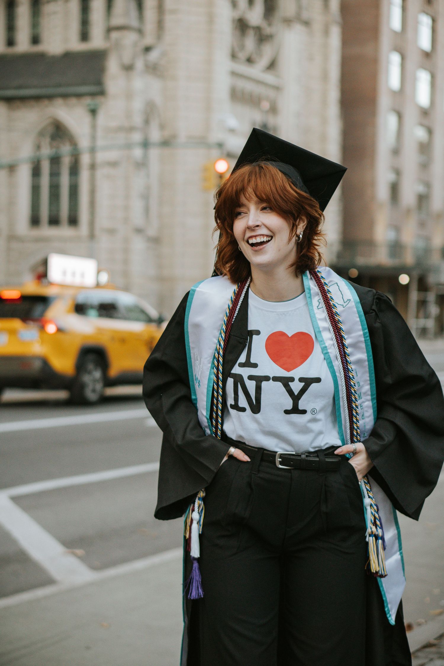 Graduate in cap and gown wearing I Love NY shirt posing on NYC street with yellow taxi visible.