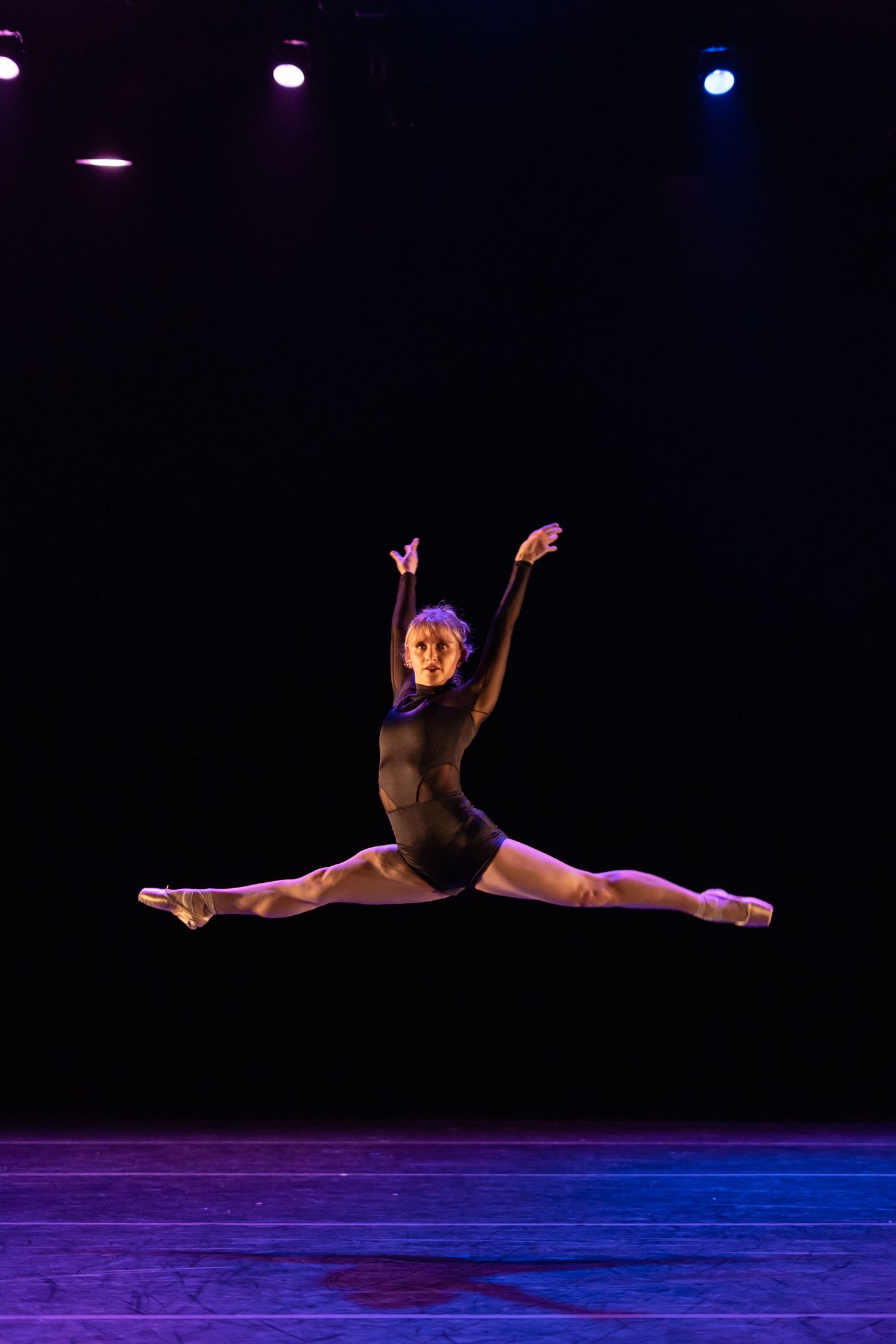 Dancer executes powerful leap across stage under purple and blue lighting.