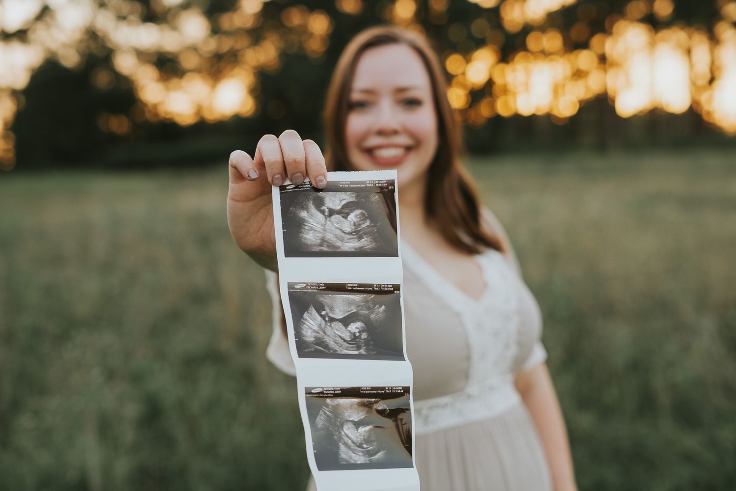 Person in white dress holding ultrasound photos during sunset maternity shoot.
