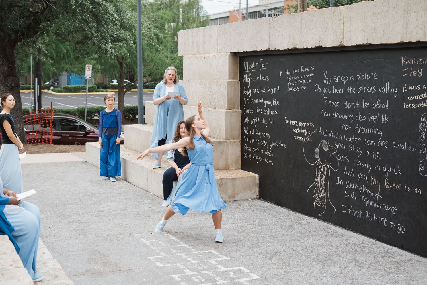 Dancers in blue dresses perform outdoors near a large chalkboard wall with handwritten text in an urban setting.