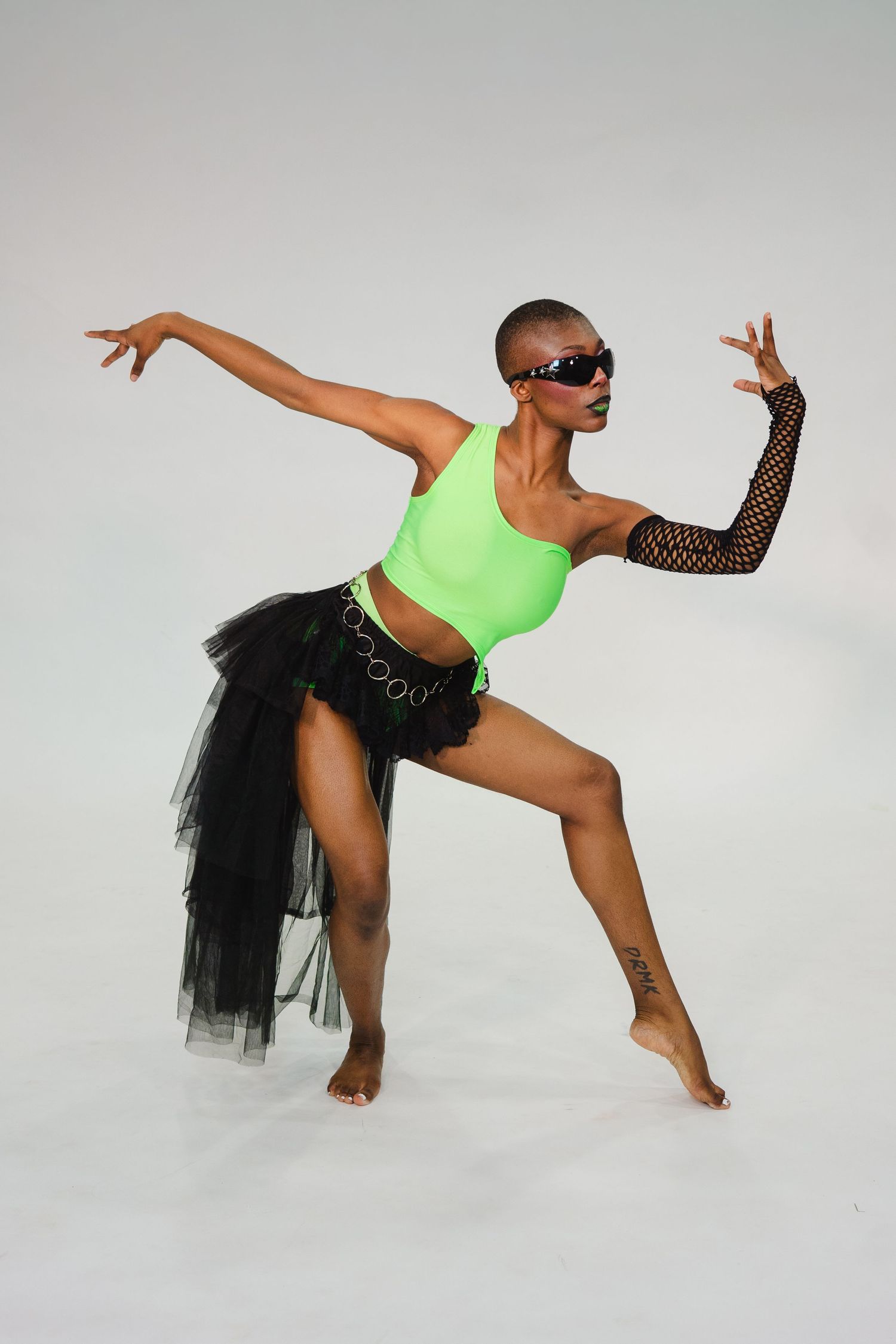 A dancer in a neon green top, black tutu skirt, fishnet gloves, and sunglasses strikes a dynamic pose on a white background.