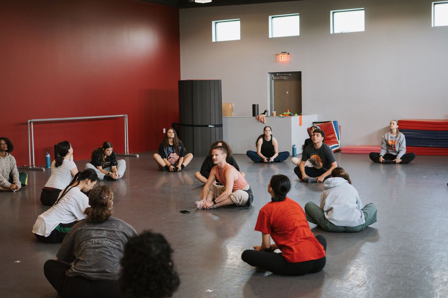 Students sit in a circle on a dance studio floor during a group discussion or class activity.
