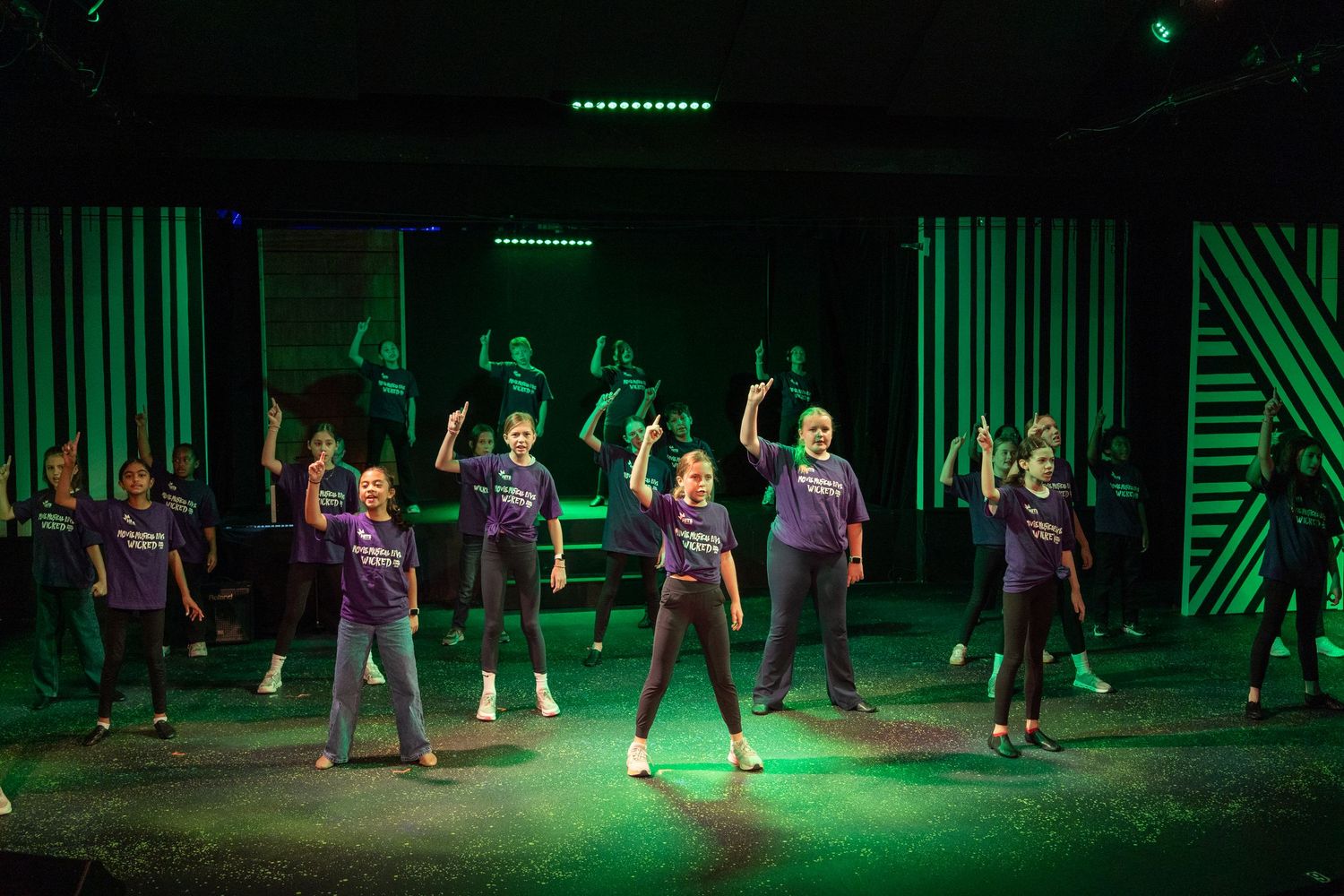 Youth performers in purple shirts dance on a green-lit stage during a theatrical group performance.