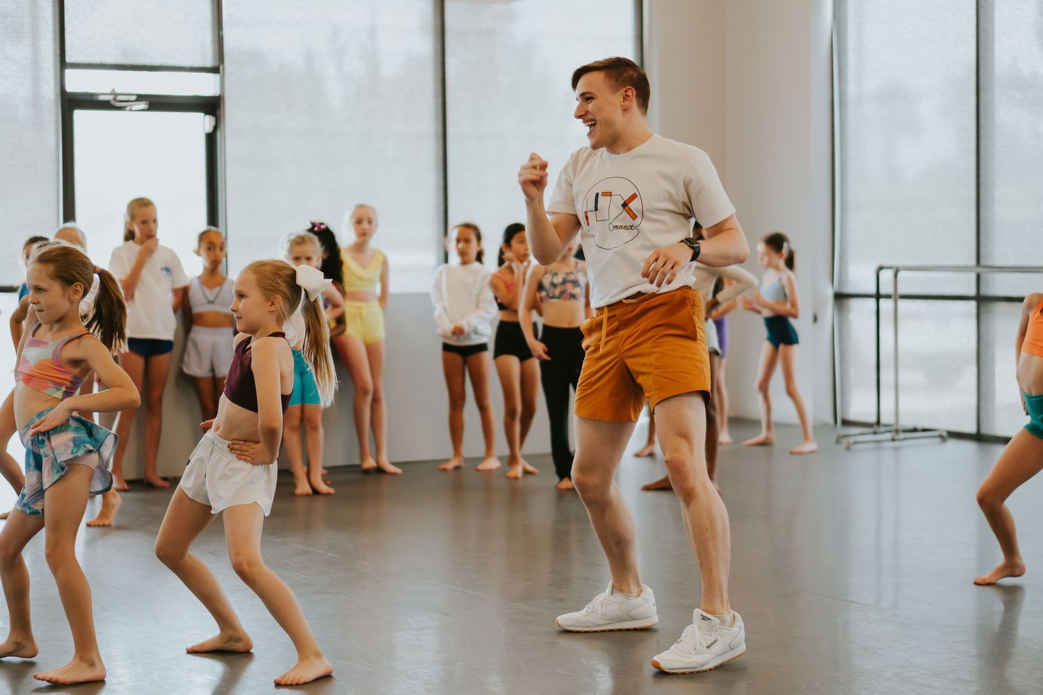 Dance students practicing dynamic movements in formation across a bright studio with white walls and windows.