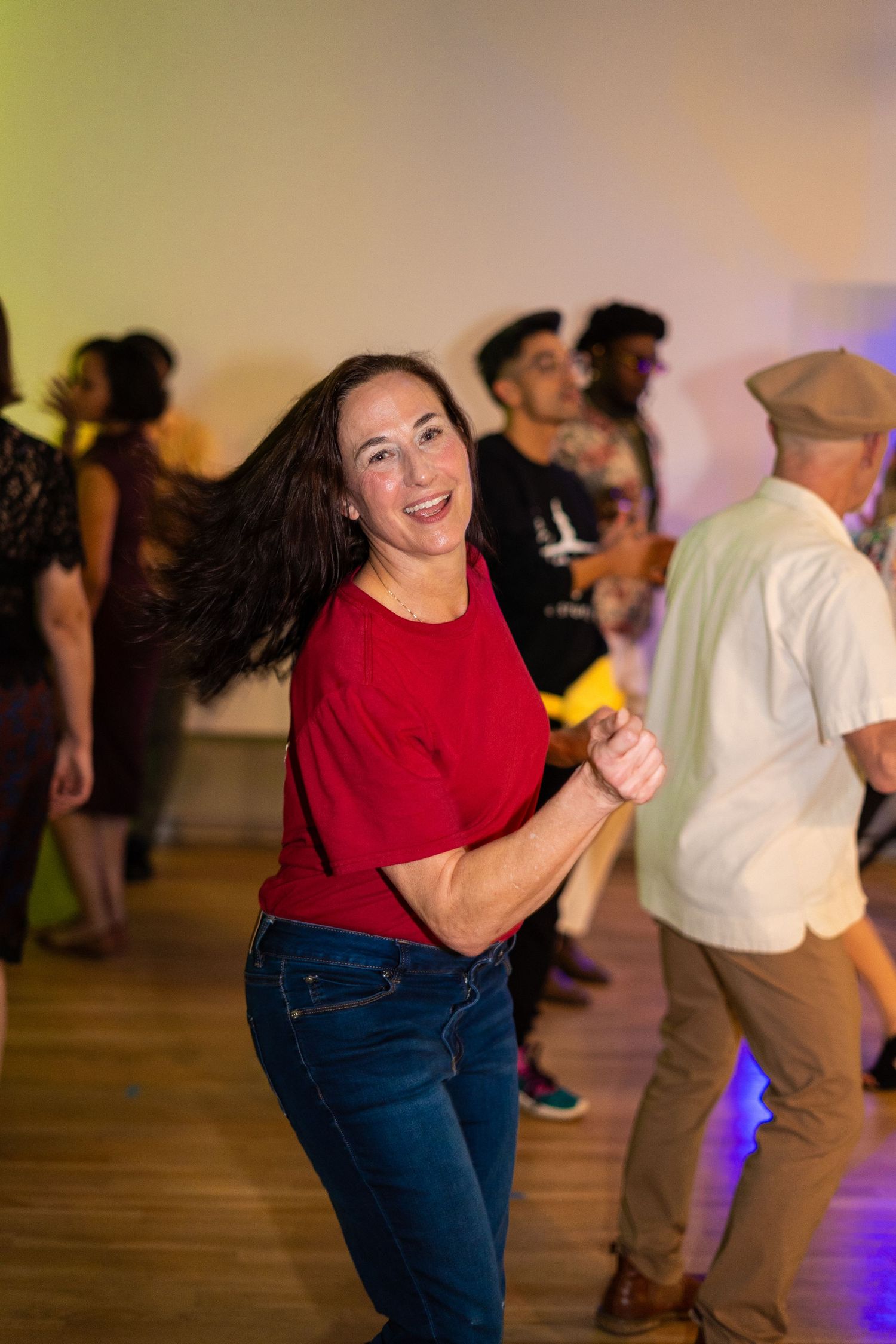 A dancer in red shirt and jeans moves energetically on a wooden dance floor at a social event.