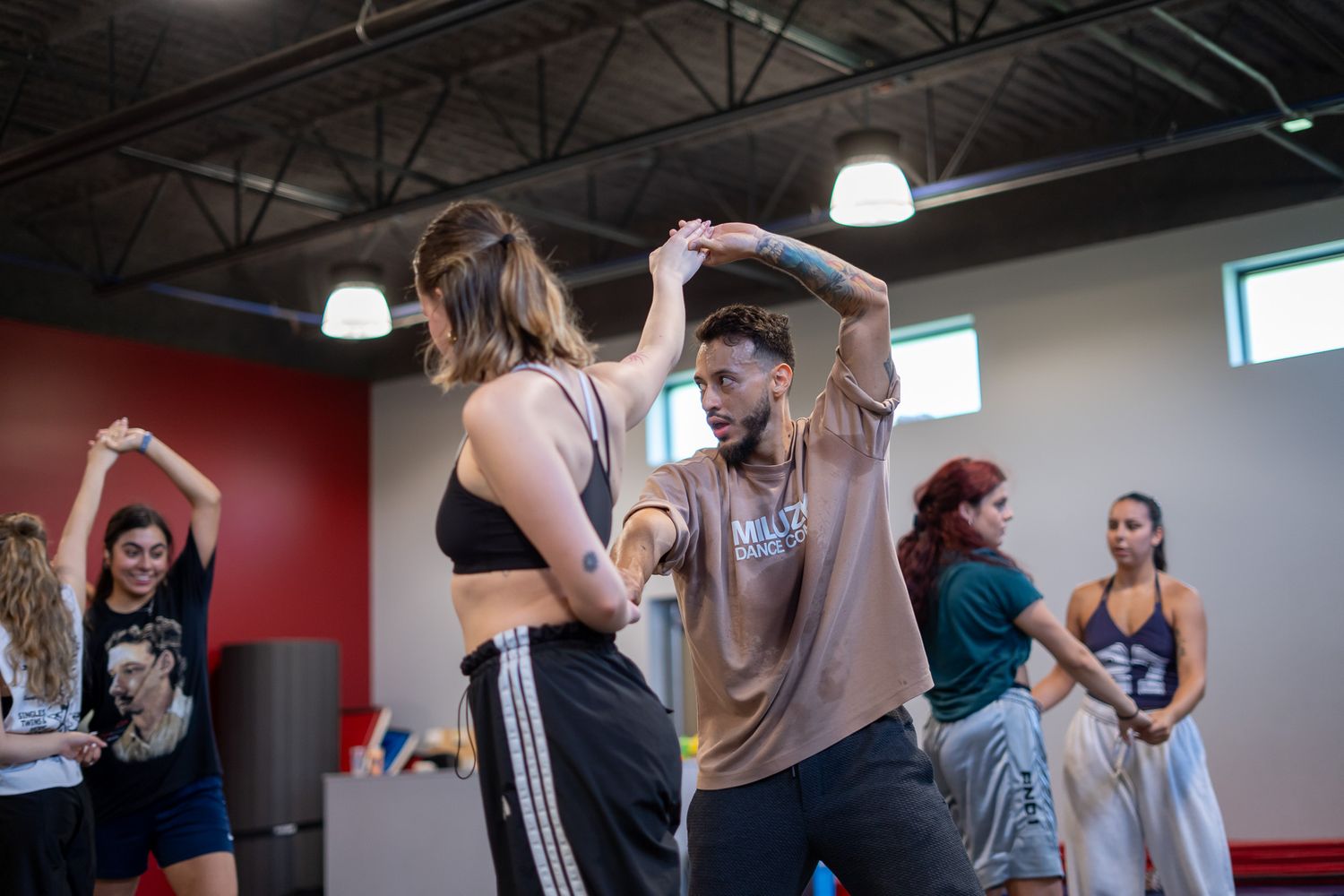 Instructor guides student in dance class with other dancers practicing in a modern studio.