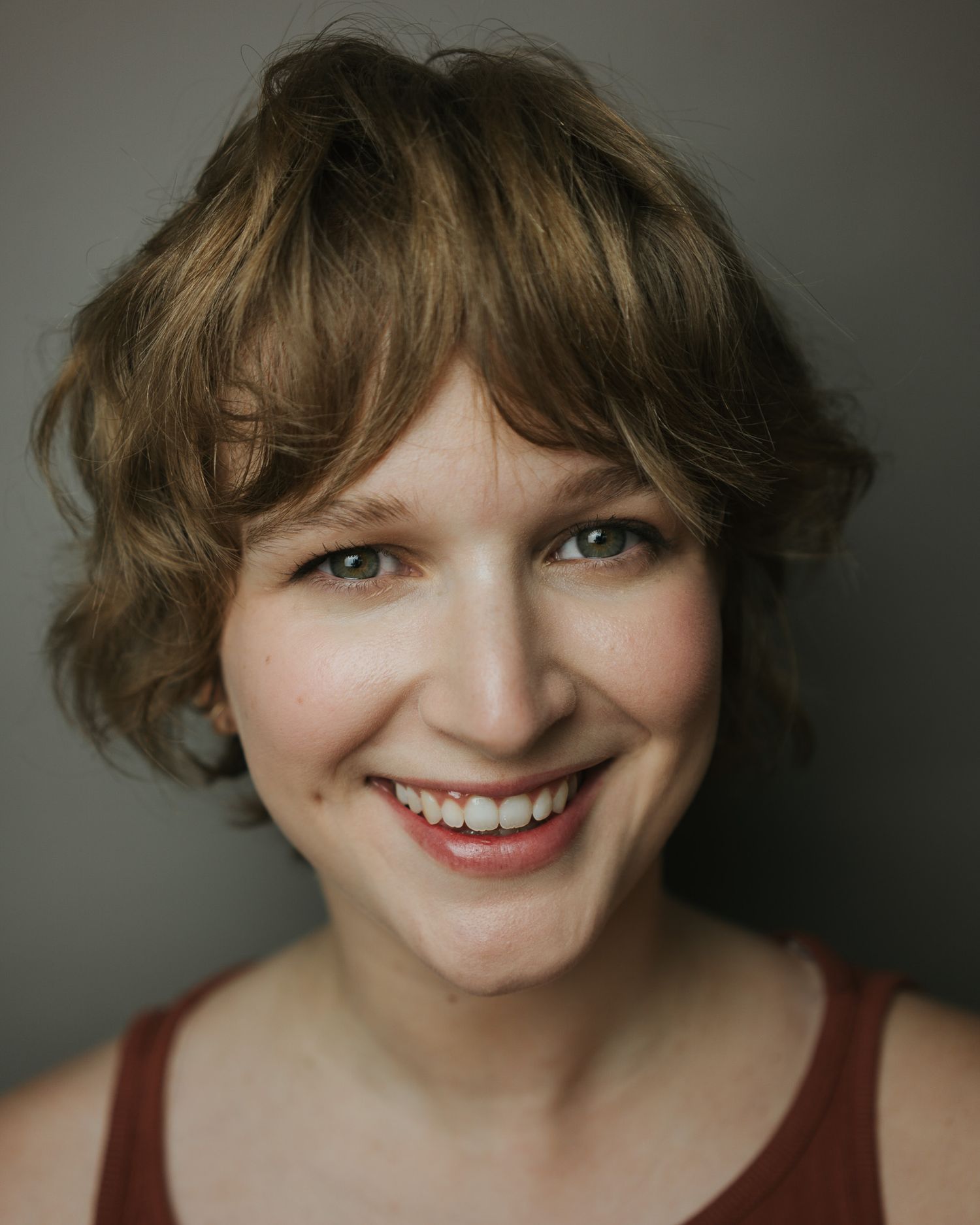 Portrait of someone with short wavy brown hair, bright eyes and a warm genuine smile wearing a burgundy top.